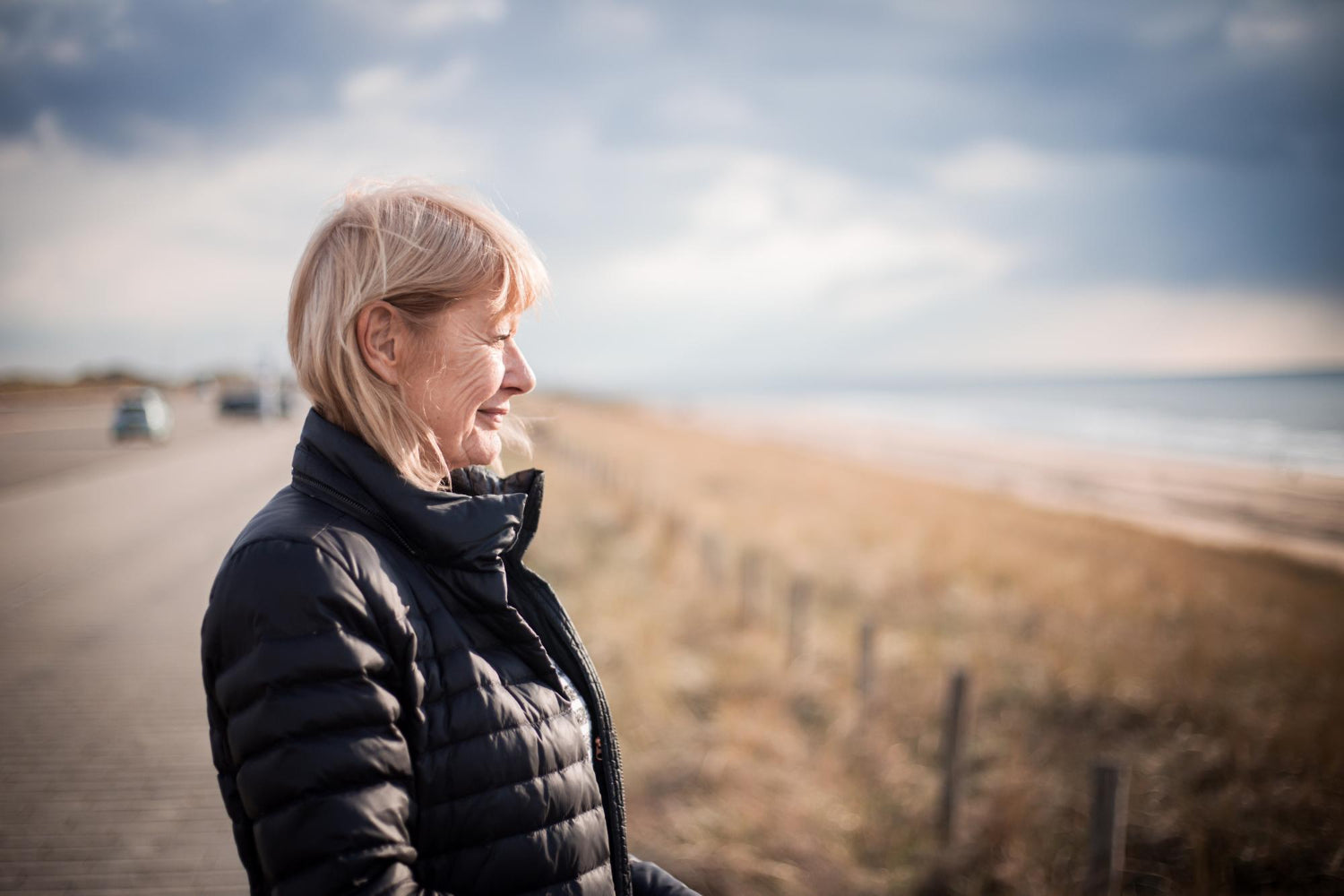 Frau im Profil bei einem Spaziergang am Meer, windiges Wetter und weiche Küstenlandschaft im Hintergrund