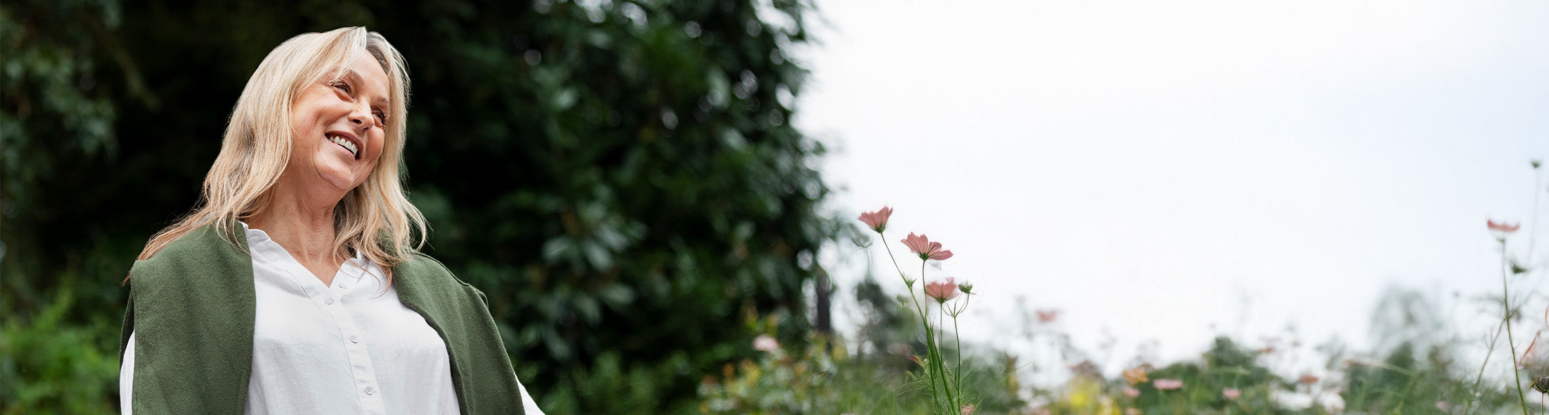 Frau lächelnd in einem Garten, umgeben von Pflanzen und Blumen, helle natürliche Umgebung