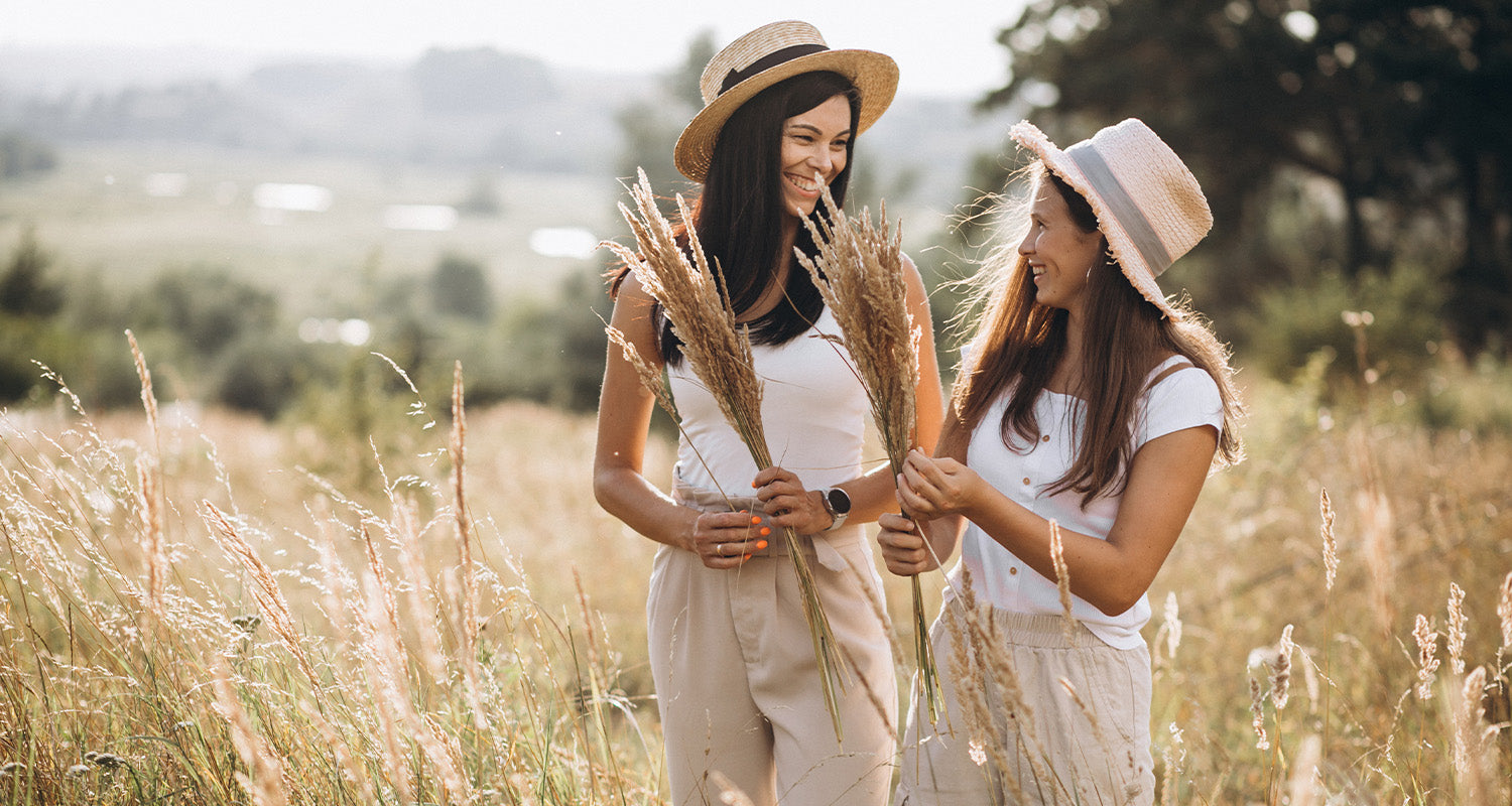 Zwei Frauen stehen lachend in einer sonnigen Wiese und halten getrocknete Gräser in den Händen